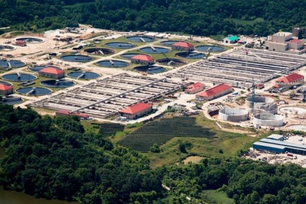 Aerial view of a wastewater treatment plant with circular settling tanks and rectangular treatment basins surrounded by forest along a river.