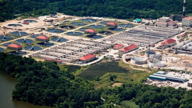 Aerial view of a wastewater treatment plant with circular settling tanks and rectangular treatment basins surrounded by forest along a river.