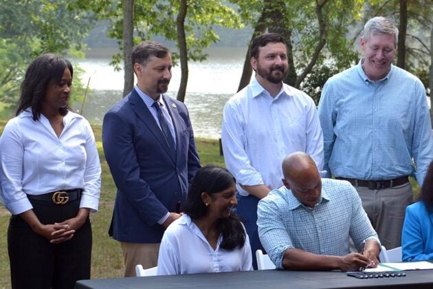 Governor Moore sits at a table signing two executive orders regarding Chesapeake Bay restoration while several people stand behind him.