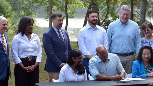 Governor Moore sits at a table signing two executive orders regarding Chesapeake Bay restoration while several people stand behind him.