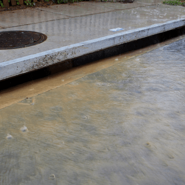 Stormwater flooding covers a street with murky water flowing into a storm drain.