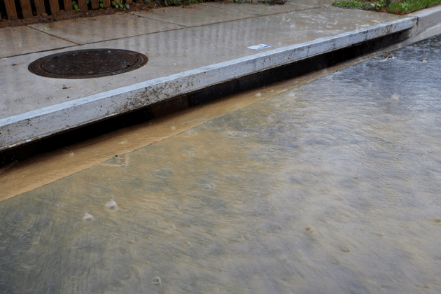 Stormwater flooding covers a street with murky water flowing into a storm drain.
