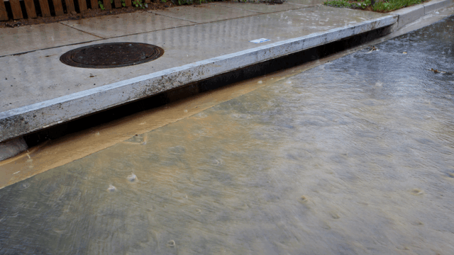 Stormwater flooding covers a street with murky water flowing into a storm drain.