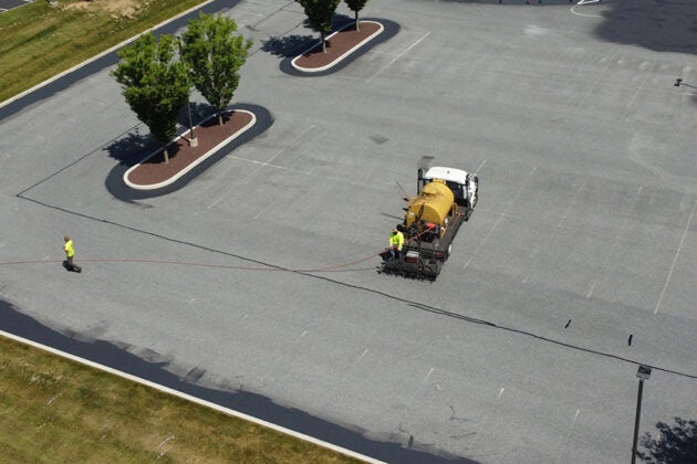 An aerial view of a sealcoating truck with a yellow tank applying pavement sealant in a parking lot with landscaped tree islands