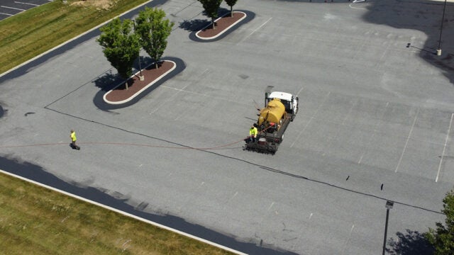 An aerial view of a sealcoating truck with a yellow tank applying pavement sealant in a parking lot with landscaped tree islands.