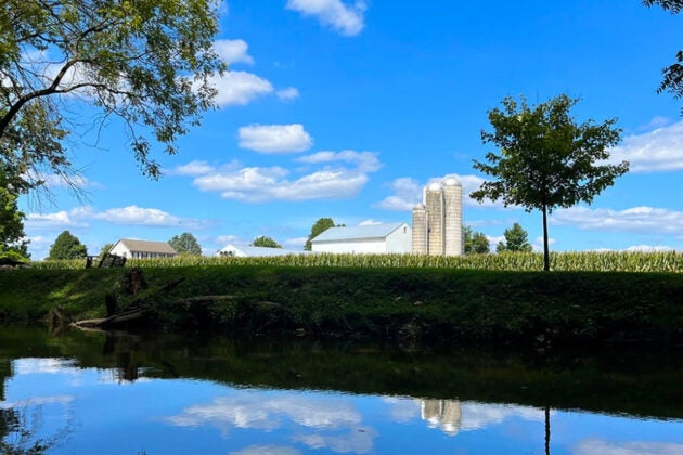 A farm field with a farm building and silos in the distance slopes down to a river.