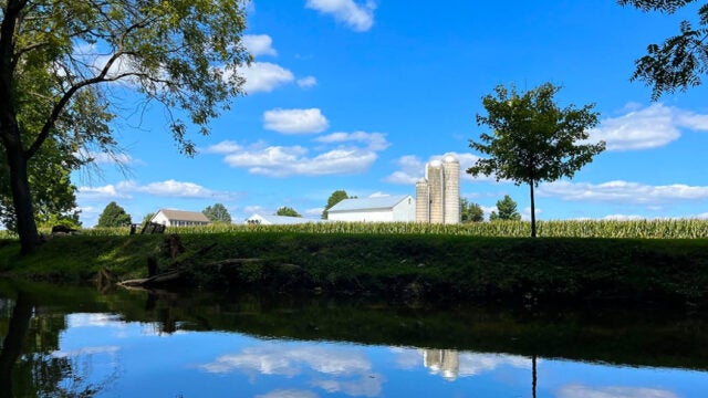 A farm field with a farm building and silos in the distance slopes down to a river.