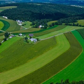 An aerial view shows contoured farm fields in varying shades of green rolling across the landscape with forests and farmhouses interspersed throughout the land