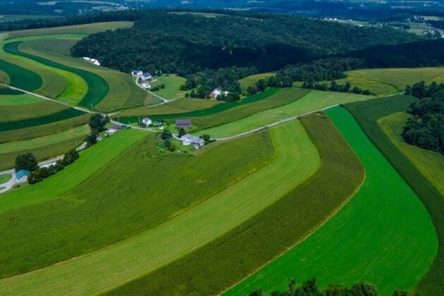 An aerial view shows contoured farm fields in varying shades of green rolling across the landscape with forests and farmhouses interspersed throughout the land