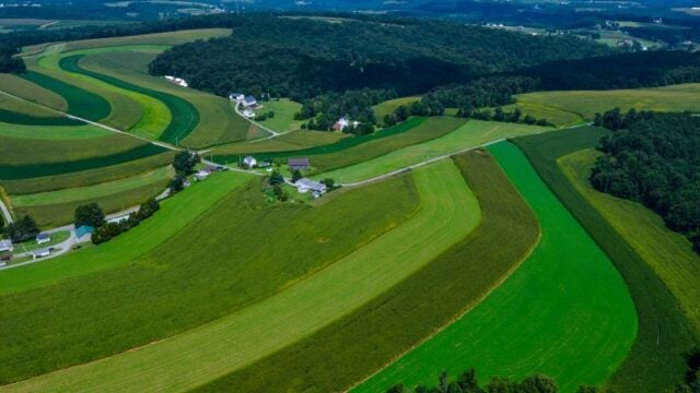 An aerial view shows contoured farm fields in varying shades of green rolling across the landscape with forests and farmhouses interspersed throughout the land.