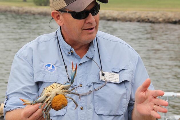 A close-up of Chris Moore, wearing a baseball hat and sunglasses, and holding a crab.