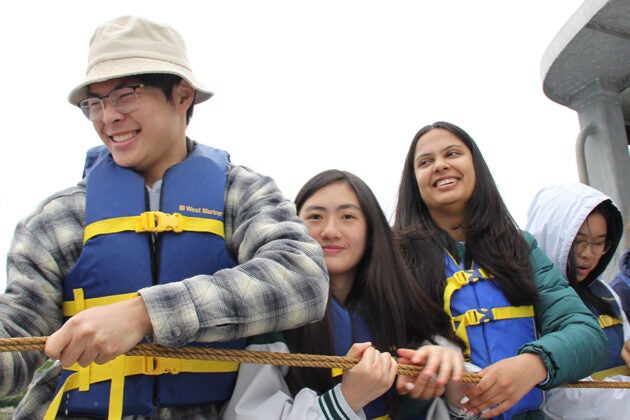 Students in blue life jackets haul on a rope aboard a boat.