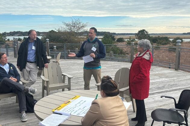 A group of people hold a discussion on an outdoor deck overlooking the marsh.
