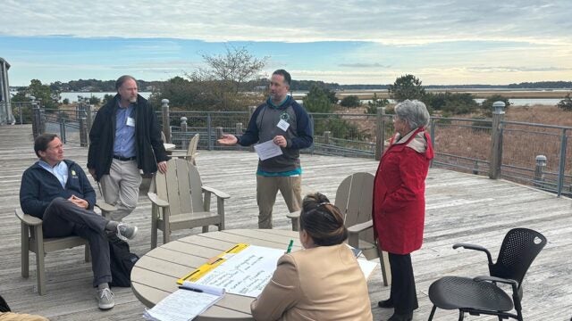 A group of people hold a discussion on an outdoor deck overlooking the marsh.