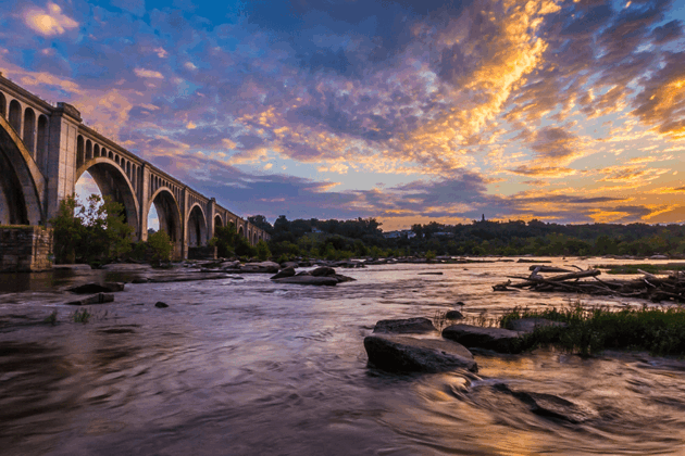 A stone arch bridge crosses the James River at sunset, with dramatic golden clouds and rocky water below.