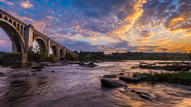 A stone arch bridge crosses the James River at sunset, with dramatic golden clouds and rocky water below.