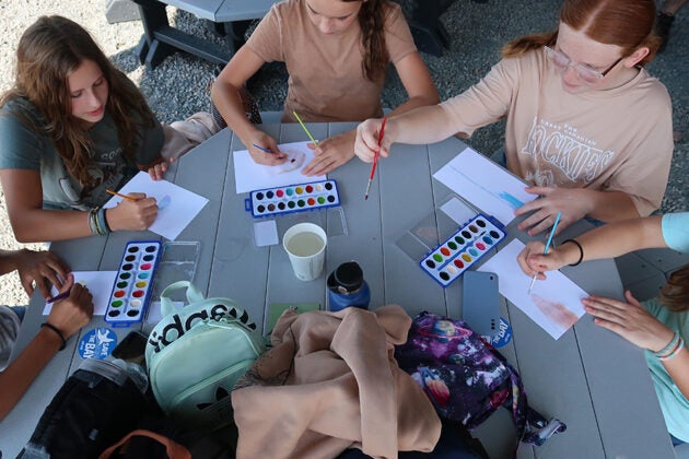 Students sit around a table using watercolors to paint on white paper.