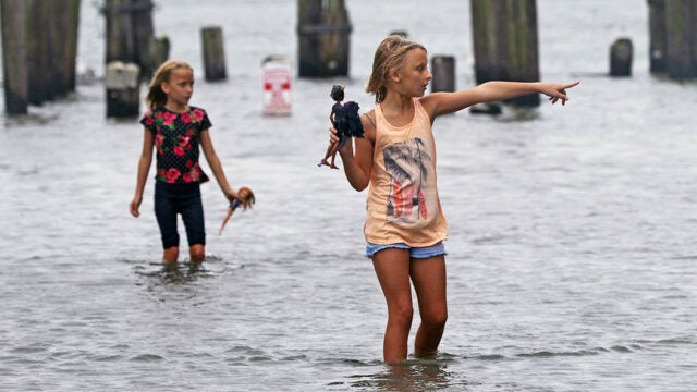 Two young girls holding dolls stand in floodwaters.