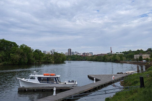 A white boat docked at a wooden pier on the James River with the Richmond skyline visible in the distance under cloudy skies.