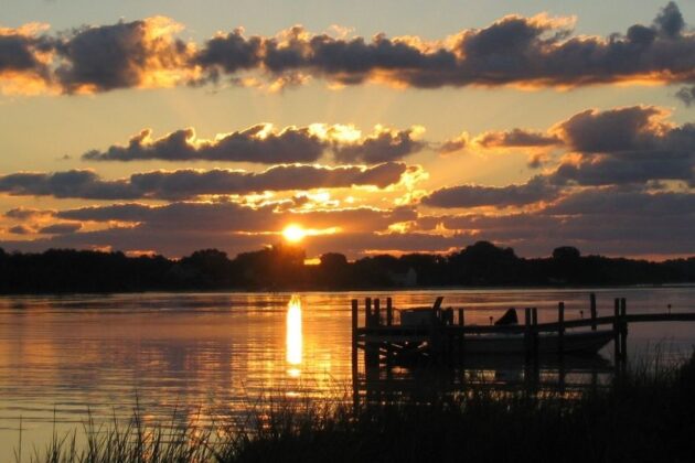 Sunrise over the Chesapeake Bay with dramatic clouds, golden light reflecting on calm water, and a silhouetted dock in the foreground.