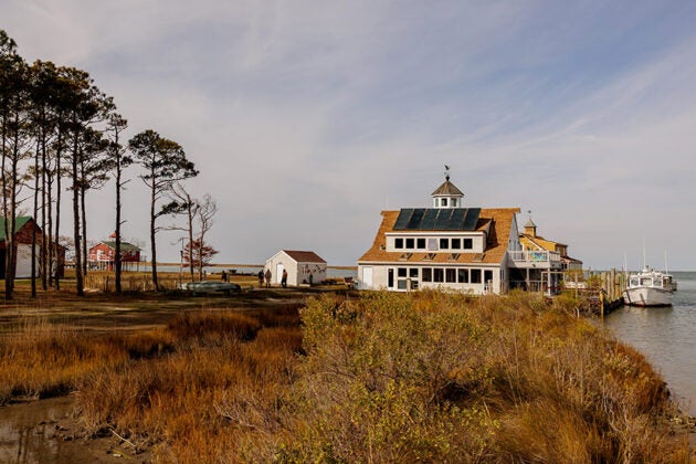 The Port Isobel Education Center stands along the waterfront surrounded by golden marsh grasses, trees and boats visible at the dock.