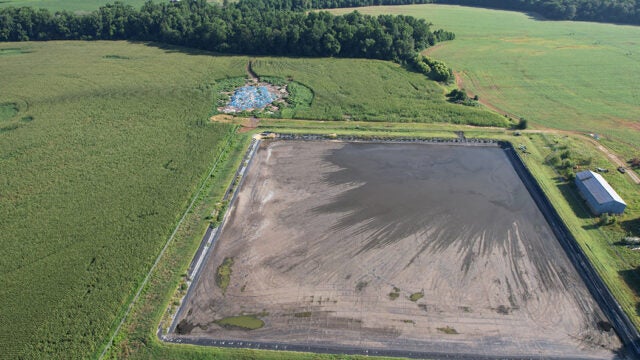 Aerial view of a large rectangular wastewater lagoon surrounded by green agricultural fields and farm buildings.
