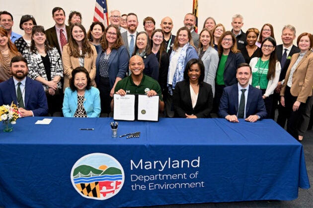 Governor Moore displays signed outdoor learning legislation at the Maryland Department of the Environment table surrounded by officials and supporters.