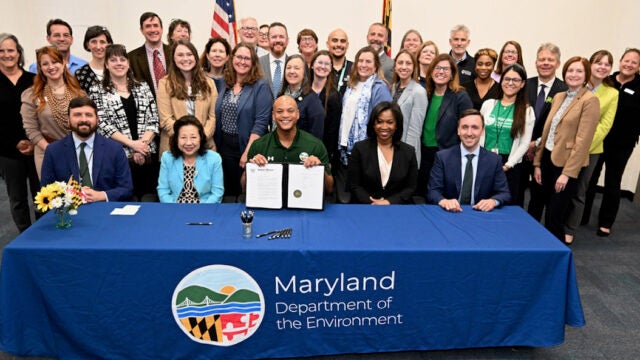 Governor Moore displays signed outdoor learning legislation at the Maryland Department of the Environment table surrounded by officials and supporters.