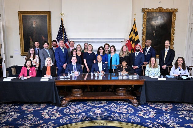 Governor Moore sits at a ceremonial desk surrounded by officials and supporters during a bill signing ceremony at the Maryland State House.