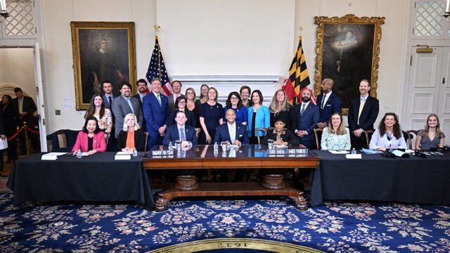 Governor Moore sits at a ceremonial desk surrounded by officials and supporters during a bill signing ceremony at the Maryland State House.