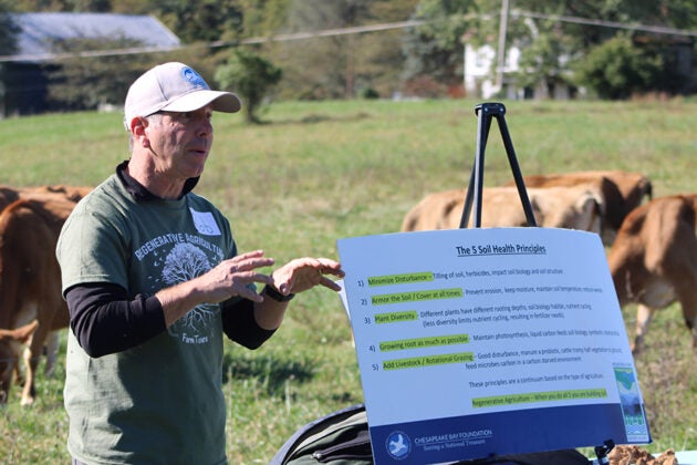A man presents about the five soil health principles using a display board, with cattle grazing in a pasture behind him.