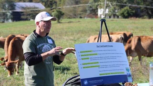 A man presents about the five soil health principles using a display board, with cattle grazing in a pasture behind him.